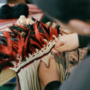 Hands weaving a korowai made of black and red feathers.