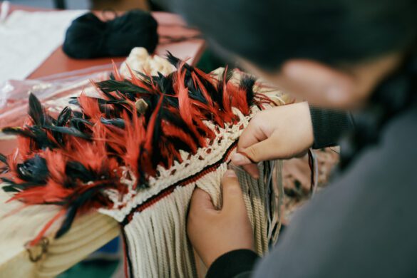 Hands weaving a korowai made of black and red feathers.