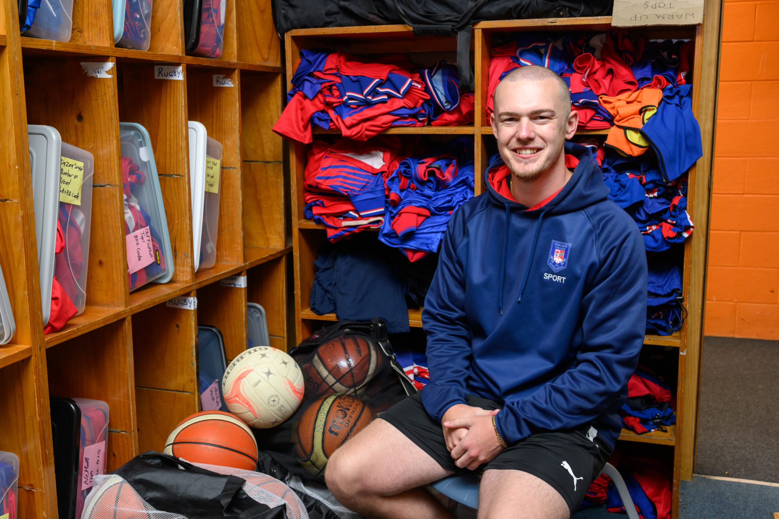 Smiling Jayden Robson sitting in small room full of sports uniforms and equipment.